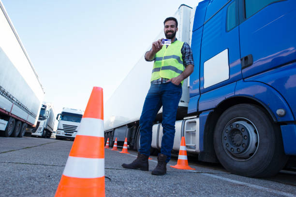 Portrait of middle aged bearded truck driver standing by his truck and showing his commercial driver license. Truck driving school and job openings. For educational purposes only