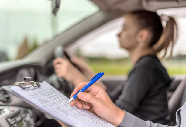 Teenage Girl Taking A Driving Lesson during bright sunny day. Closeup of a instructor man writing on a clipboard and taking notes while sitting next to teen driver. For educational purposes only
